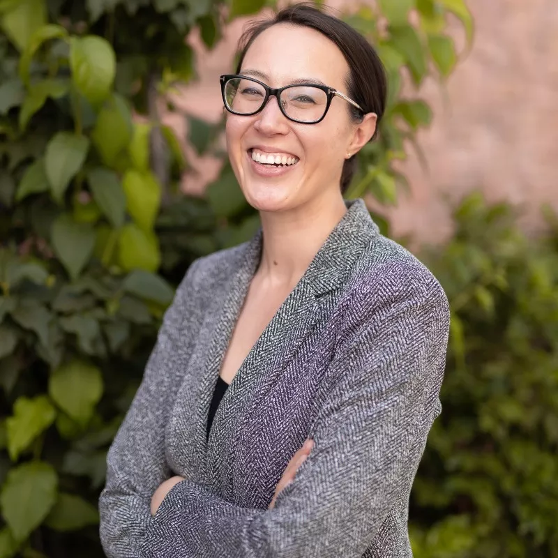 Headshot of photo of smiling human in a knit black and white blazer with arms folder. The backdrop is green ivy. The headshot is of Jaimie Shaff with black eye glasses and hair in a low ponytail.